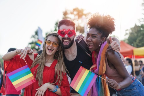 Three friends celebrate at a pride event, holding rainbow flags and wearing colorful accessories, smiling joyfully.