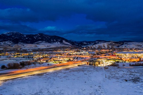 A snowy landscape at dusk, with a town illuminated by lights and mountains in the background under a cloudy sky.