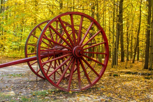 A large red wooden wheel rests on a forest path surrounded by vibrant autumn foliage.