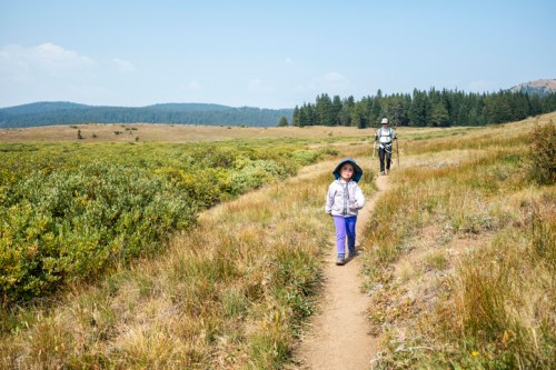 A child and an adult walk along a grassy trail in a scenic landscape with trees and mountains in the background.