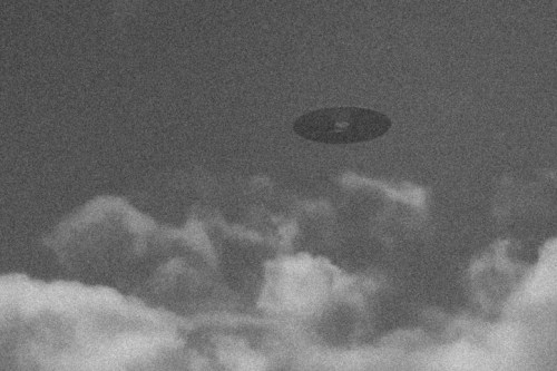 A dark, unidentified flying object hovering above fluffy clouds in a grayscale sky.