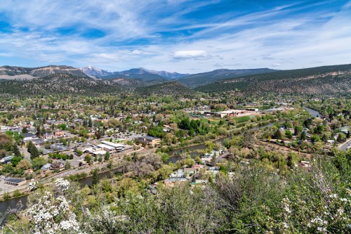 A scenic view of a valley with a river, surrounded by mountains and greenery under a blue sky with clouds.