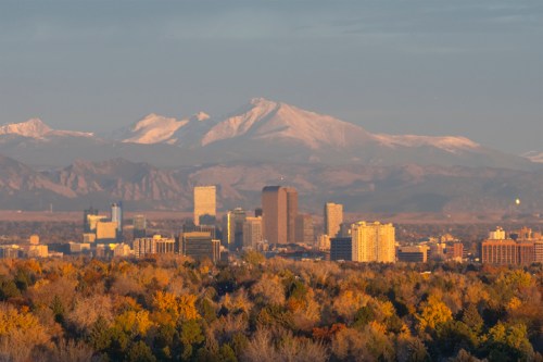 A city skyline with autumn trees in the foreground and snow-capped mountains in the background under a clear sky.