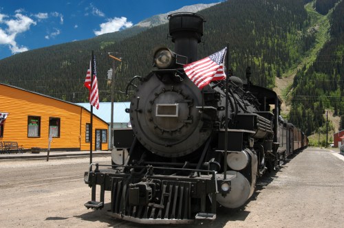 A vintage steam locomotive with American flags, set against a backdrop of mountains and a colorful building.