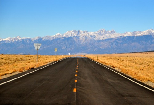A straight road stretches into the distance, flanked by golden grass and snow-capped mountains under a clear blue sky.