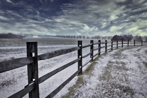 A snowy landscape with a wooden fence lining a path, under a dramatic sky with clouds.