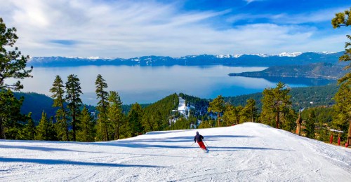 A skier descends a snowy slope with a panoramic view of Lake Tahoe and mountains in the background.