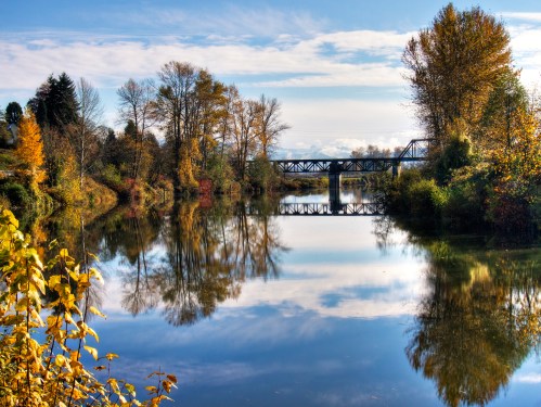 A serene river scene with autumn trees reflecting in the water and a bridge in the background under a blue sky.