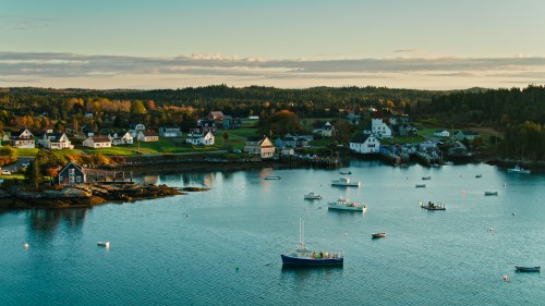A serene coastal village with boats in the water, surrounded by trees and houses under a clear sky.