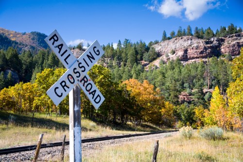 A railroad crossing sign stands in a scenic landscape with trees and mountains under a blue sky.