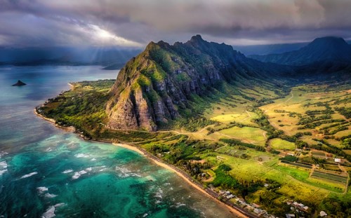 Aerial view of lush green mountains meeting a turquoise coastline under a dramatic sky.