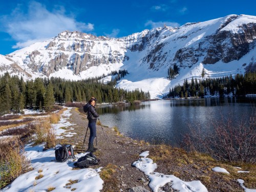 A hiker stands by a snowy lake surrounded by mountains and evergreen trees, holding a trekking pole.