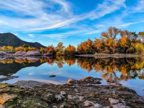 A serene landscape featuring a calm lake reflecting autumn trees and a blue sky with wispy clouds.