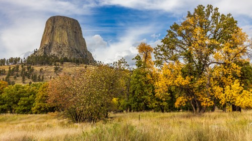 A towering rock formation surrounded by autumn trees and grassy fields under a cloudy blue sky.