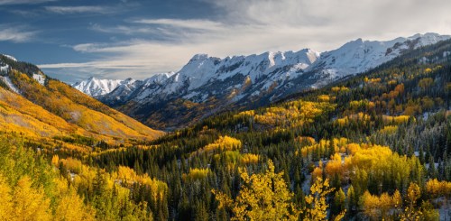 A panoramic view of snow-capped mountains surrounded by vibrant autumn foliage in a valley.