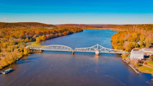 Aerial view of a river with a bridge, surrounded by autumn foliage and rolling hills under a clear blue sky.