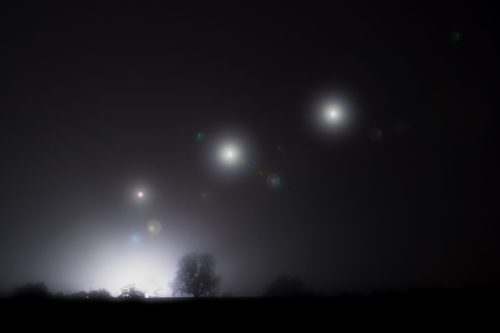 A foggy night scene with glowing orbs in the sky and a silhouette of a tree against a bright light in the background.