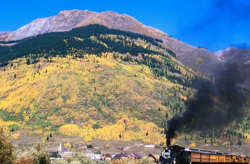 A vintage steam train travels through a vibrant autumn landscape with colorful trees and mountains in the background.
