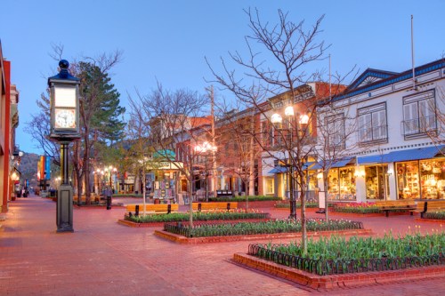 A quiet, tree-lined street at dusk, featuring shops, benches, and blooming flowers in a charming town square.