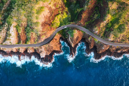 Aerial view of a winding road along a rugged coastline with cliffs and crashing waves below.