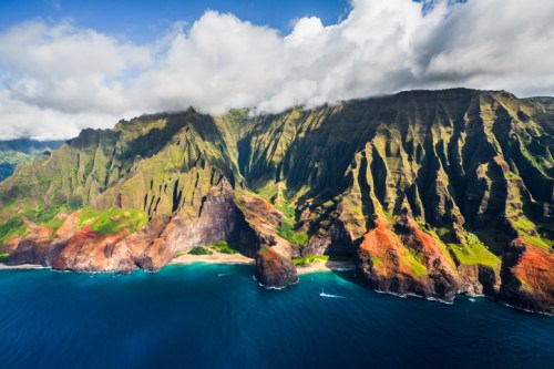 Aerial view of dramatic cliffs and lush green mountains meeting a vibrant blue ocean under a partly cloudy sky.