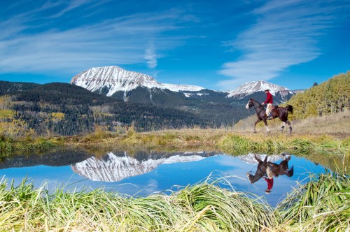 A rider on horseback walks by a serene pond, reflecting snow-capped mountains and a clear blue sky.