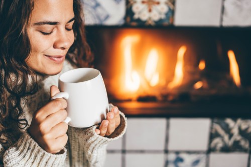 A woman with curly hair smiles while holding a mug, sitting near a cozy fireplace.