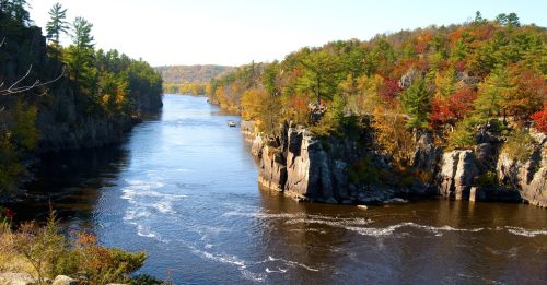 A serene river flows between rocky cliffs, surrounded by vibrant autumn foliage and clear blue skies.