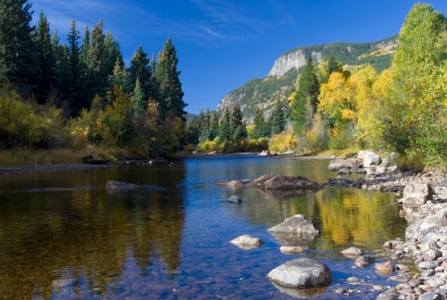 A serene river flows through a forested landscape, with colorful autumn trees and rocky banks under a clear blue sky.