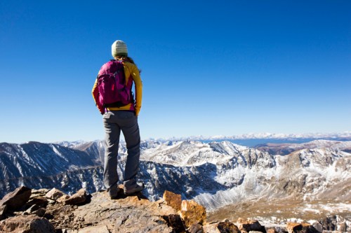 A hiker with a pink backpack stands on a rocky peak, gazing at a vast mountain landscape under a clear blue sky.