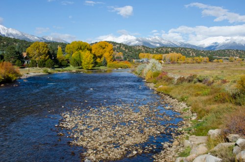 A serene river flows through a landscape of autumn trees and mountains under a clear blue sky.