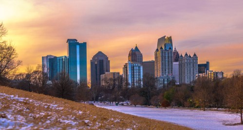 City skyline at sunset with tall buildings, a snowy park in the foreground, and a colorful sky.