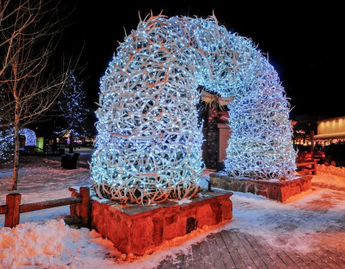 A large arch made of illuminated antlers, surrounded by snow and festive lights at night.