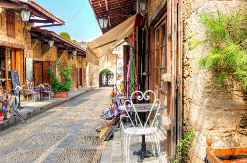 Charming cobblestone street lined with shops and a small table outside, featuring rustic stone buildings and greenery.