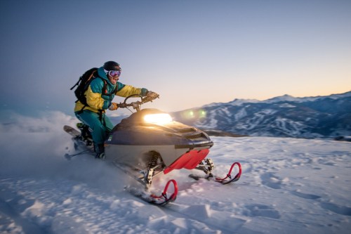 A person riding a snowmobile across a snowy landscape at sunset, with mountains in the background.