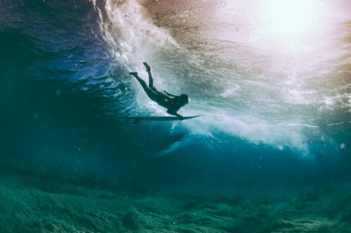 A surfer glides beneath a wave, illuminated by sunlight filtering through the water's surface.
