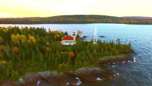 Aerial view of a lighthouse surrounded by colorful autumn trees and a calm lake, with hills in the background.