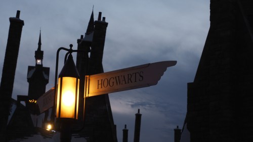 A street sign pointing to Hogwarts, illuminated by a lantern, against a twilight sky with silhouetted buildings.