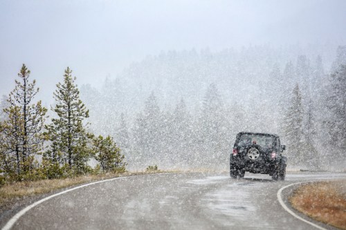 A car drives along a winding road in heavy snowfall, surrounded by trees and misty mountains.