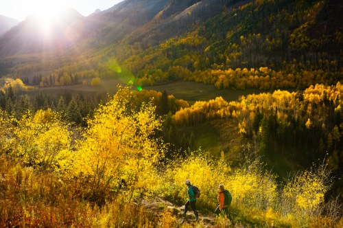 Two hikers trek along a trail surrounded by vibrant autumn foliage and mountains, with sunlight streaming through.