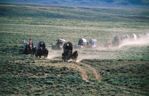 A line of covered wagons travels through a dusty, grassy landscape under a clear sky.