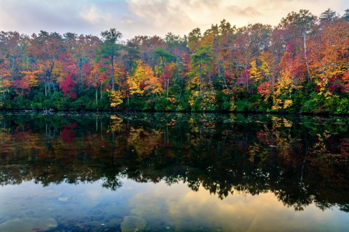 A serene lake reflects vibrant autumn foliage and a colorful sky, surrounded by trees in shades of red, orange, and green.