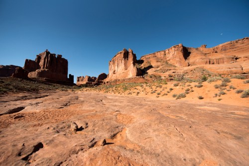 A vast desert landscape with towering rock formations under a clear blue sky. Sparse vegetation is visible on the ground.