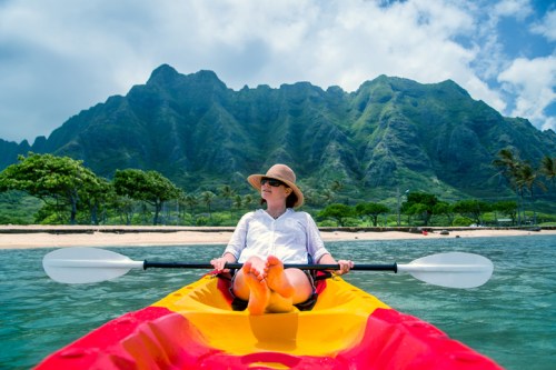 A person in a kayak relaxes on water, with lush green mountains and a sandy beach in the background.