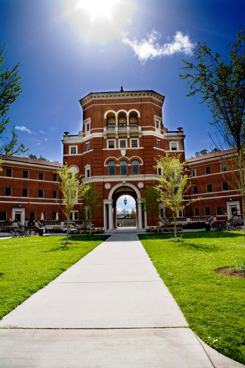A brick building with a large archway, surrounded by green grass and trees, under a bright blue sky.