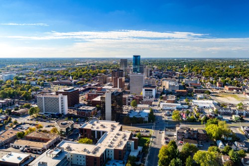 Aerial view of a city skyline with tall buildings, green trees, and a clear blue sky.