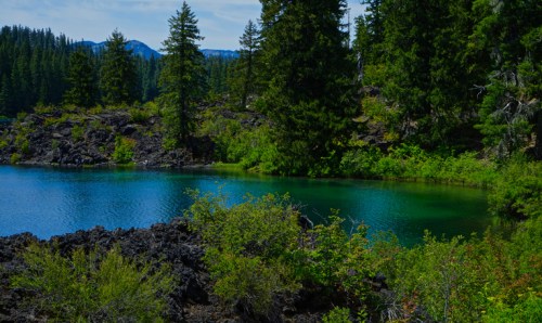 A serene lake surrounded by lush greenery and rocky terrain, with tall trees and distant mountains in the background.