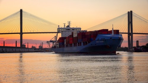 A cargo ship with colorful containers sails under a bridge at sunset, reflecting on the water.