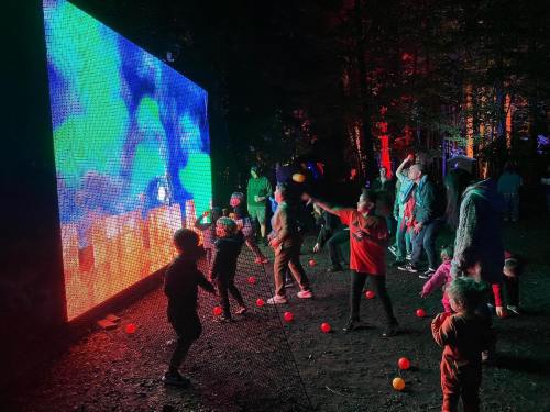 A vibrant outdoor scene with people, including children, dancing in front of a large colorful screen at night.