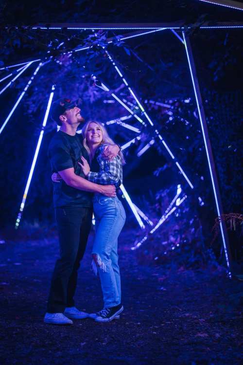 A couple embraces under colorful lights in a dark, illuminated pathway, smiling and enjoying the moment together.